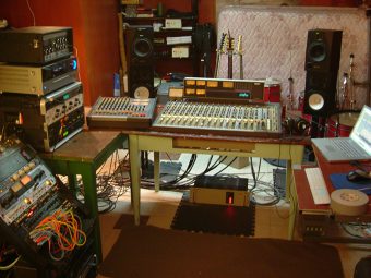 A basement with two tables full of electronic music equipment, a third table with a laptop, a pair of speakers, electric guitars, a drum kit and a mattress resting against the wall at the back