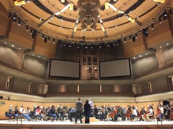 The Toronto Symphony Orchestra rehearsing onstage with two projection screens overhead, and Eliot Britton and Sandra Laronde observing from front of the stage