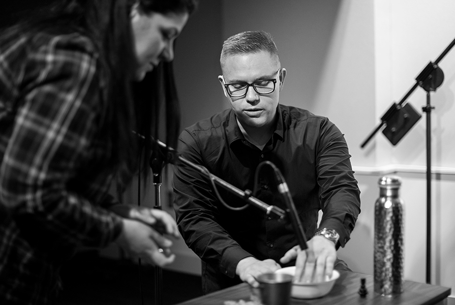 Eliot Britton sitting with his hand in a small bowl on a table underneath a microphone; a student stands slightly bent over beside him looking at the bowl