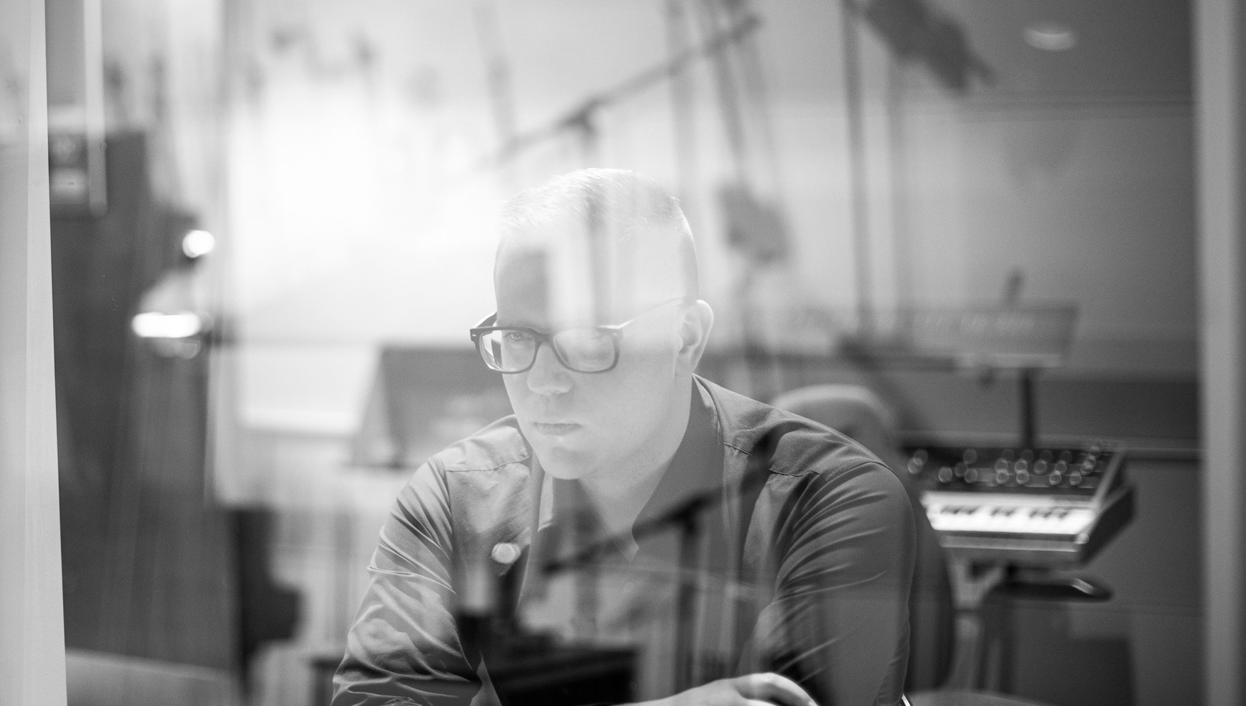 Black and white photo of Eliot Britton sitting in a music studio behind glass that is reflecting rod-like shapes