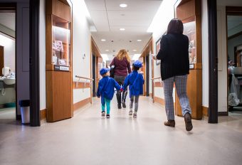 Two young children in blue uniform holding hands and walking behind and in front of two women down a hallway