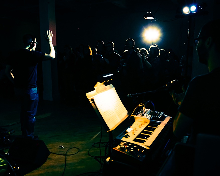 Eliot Britton standing in spotlight with one arm raised in a darkly lit space, where the outline of an audience is visible in the background and a keyboard is under spotlight in the foreground