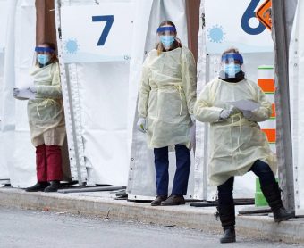 Three health-care workers wearing masks with shield and white gowns and gloves are standing outside white tents labelled "6" and "7"
