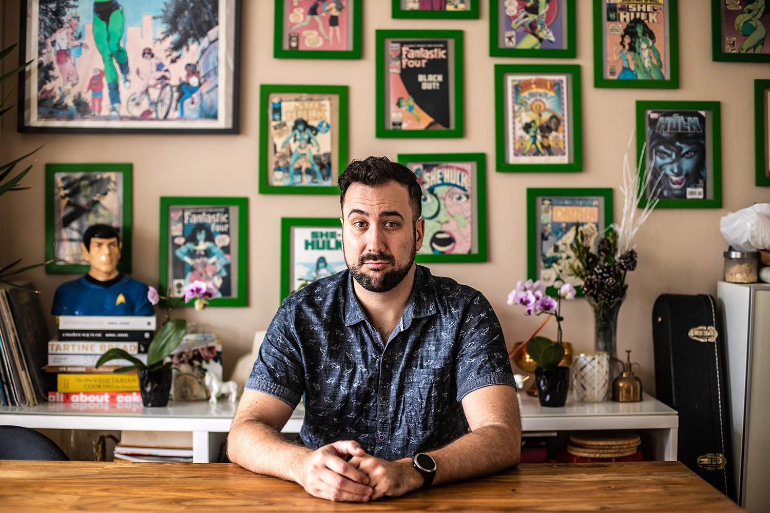 Writer and U of T grad Aaron Hagey-Mackay sits at a desk in his home. On the wall behind him hang many framed comic book covers.