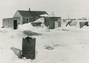Black and white photo of a house surrounded by snow-covered ground, with a steel drum barrel in the foreground.