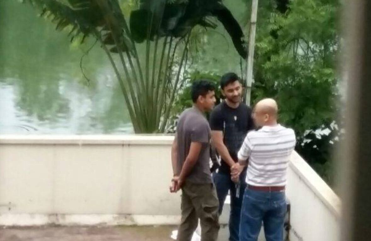 A slightly blurry photo of Tahmid Khan holding a gun and speaking with two men, whose backs are towards the camera, at the corner of a rooftop overlooking water and trees