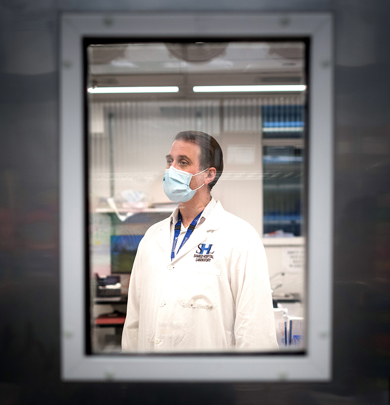 Photo of U of T and Sunnybrook Health Sciences Centre clinical microbiologist Robert Kozak wearing a lab coat and a mask, taken through a small window in a door of a lab