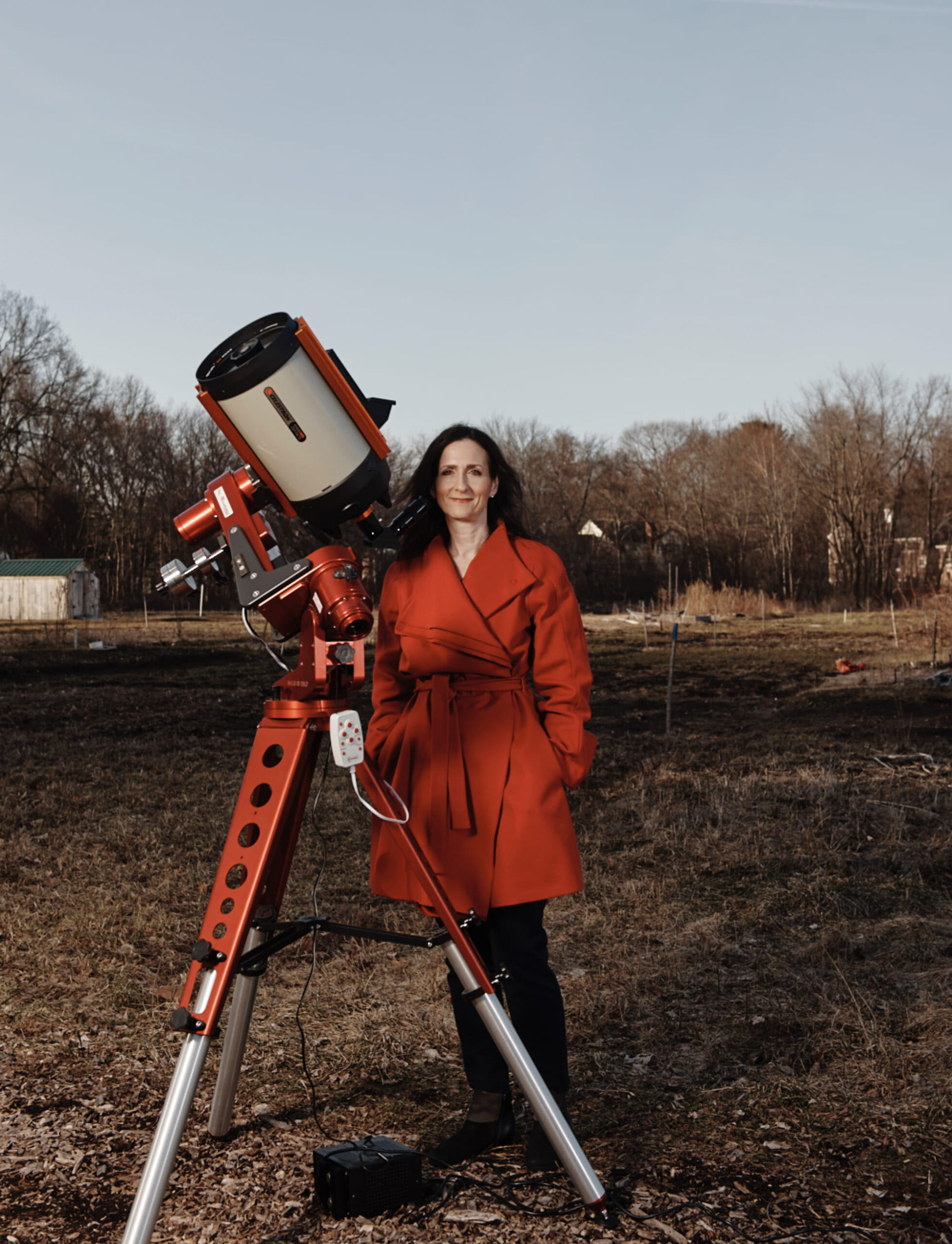 A woman with dark hair wearing a red coat stands next to a telescope in a field. The sky is blue, and there are trees in the distance behind her