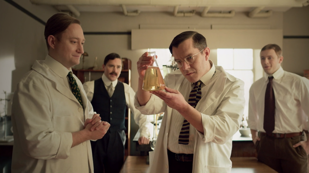 A man in a lab coat in an early 20th century laboratory inspects a beaker full of liquid as three other men look on