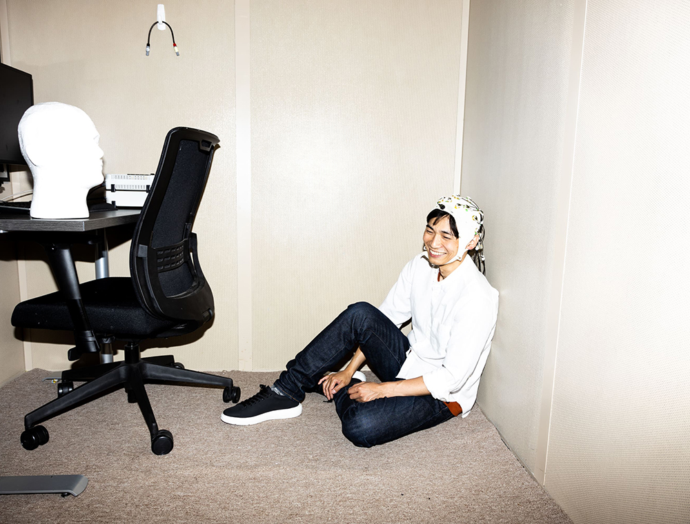 Prof. Keisuke Fukuda wearing a white cap, sitting on the ground in front of a desk and office chair