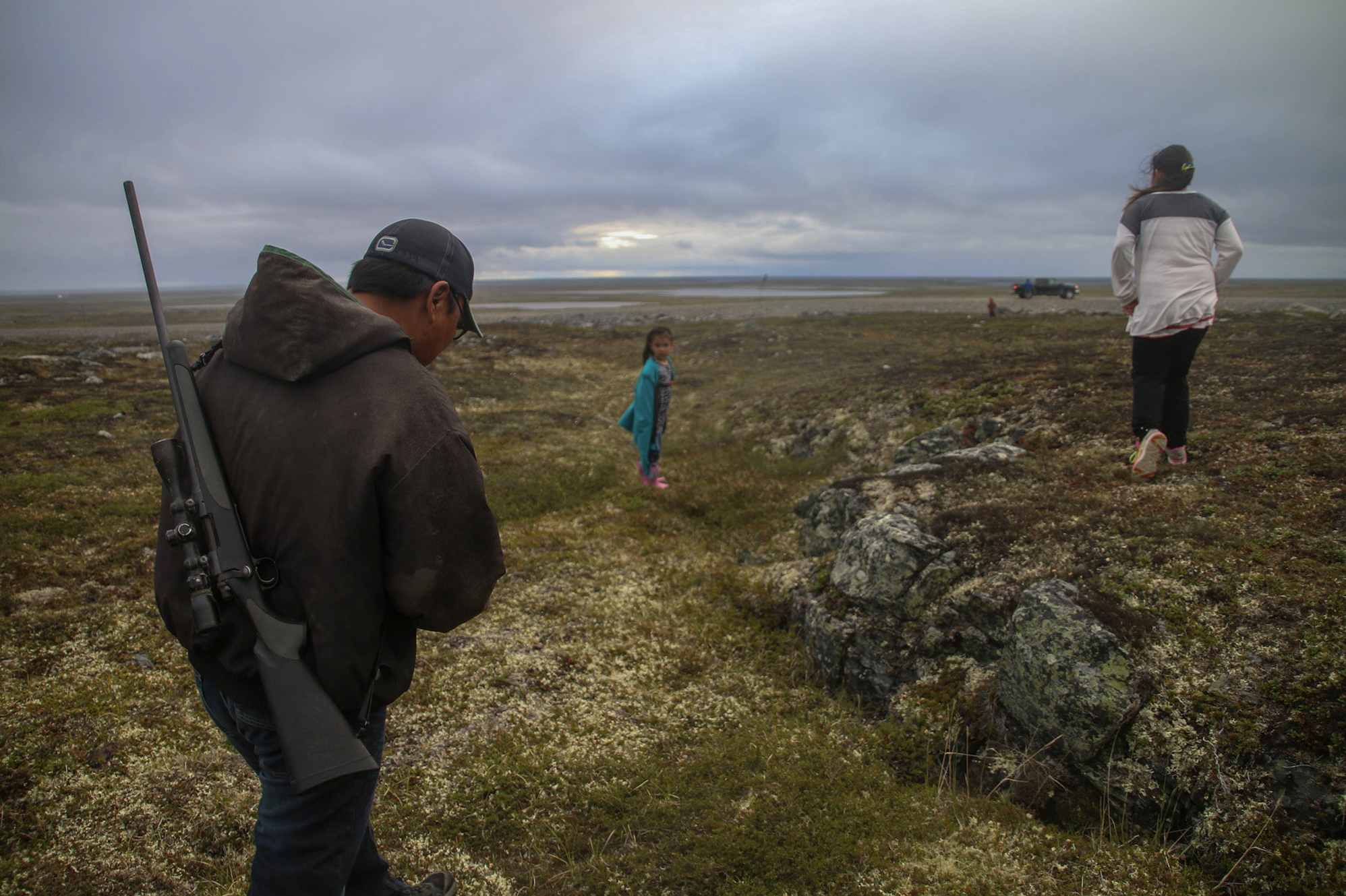 A man with a gun strapped to his back, a woman, and a young girl walking across a field toward a vehicle in the distance