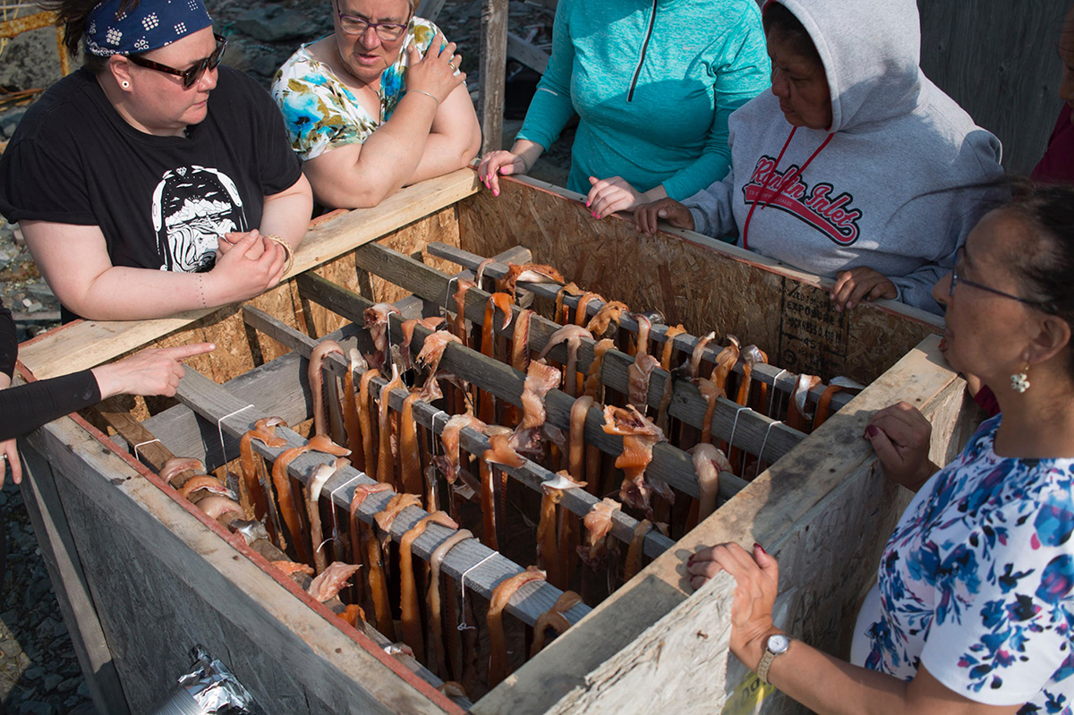 A group of women gathered around a smoker with strips of fish on racks inside