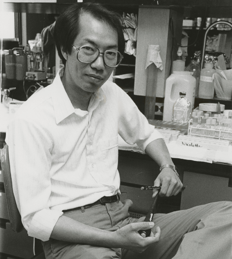 Black and white photo of Tak Mak seated in front of a lab table with vials, bottles and other equipment on top
