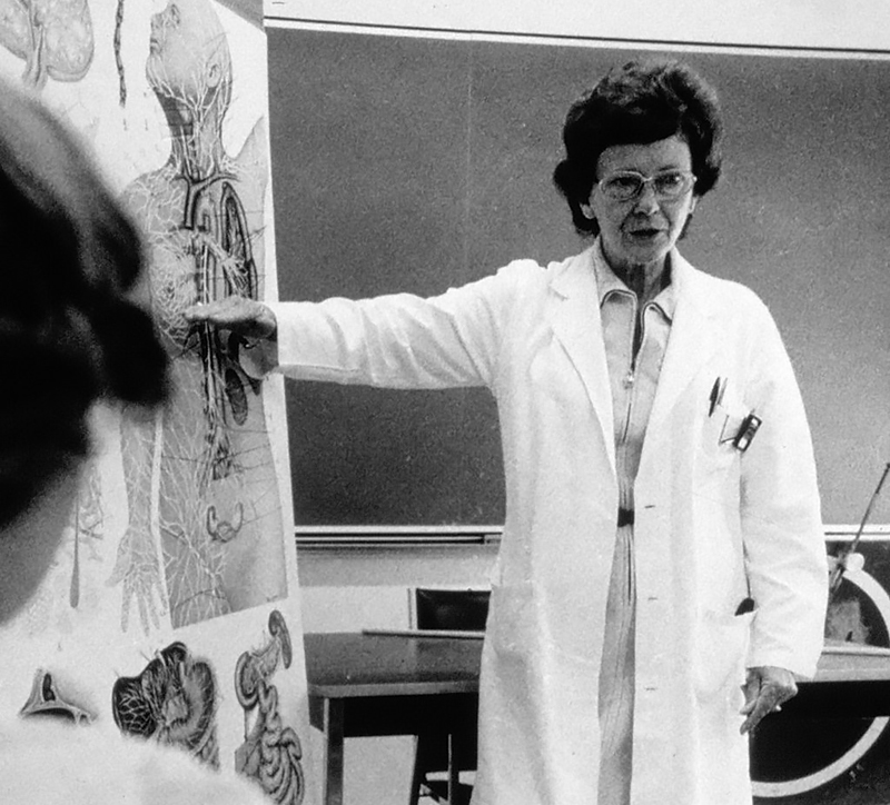Black and white photo of Vera Peters giving a lecture in a classroom, one arm gesturing toward a section of an anatomy drawing