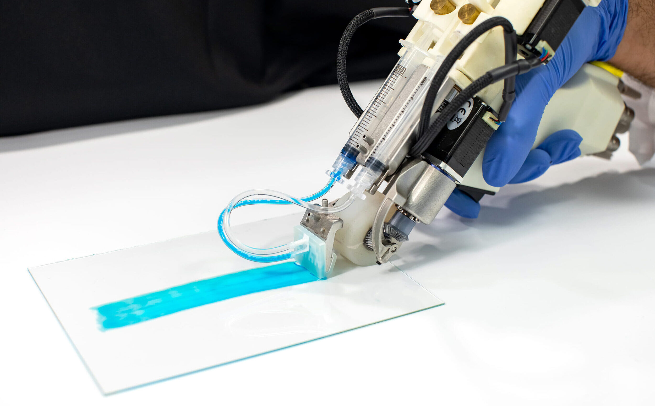 A device with a small roller applies a blue liquid along a smooth, rectangular glass surface
