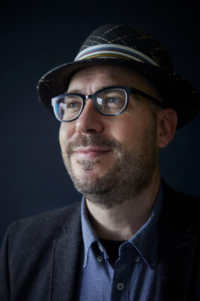 Close up of Prof. Alán Aspuru-Guzik wearing a fedora hat against a solid dark-grey background