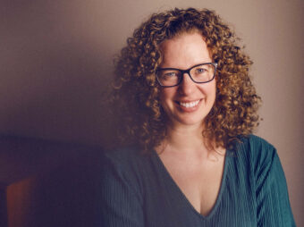 Close up of Fiona Rawle, wearing glasses and a teal ribbed sweater, smiling at the camera