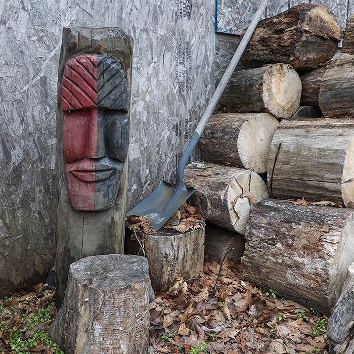 A wood carving of a face, painted half in red and half in black, next to a pile of logs