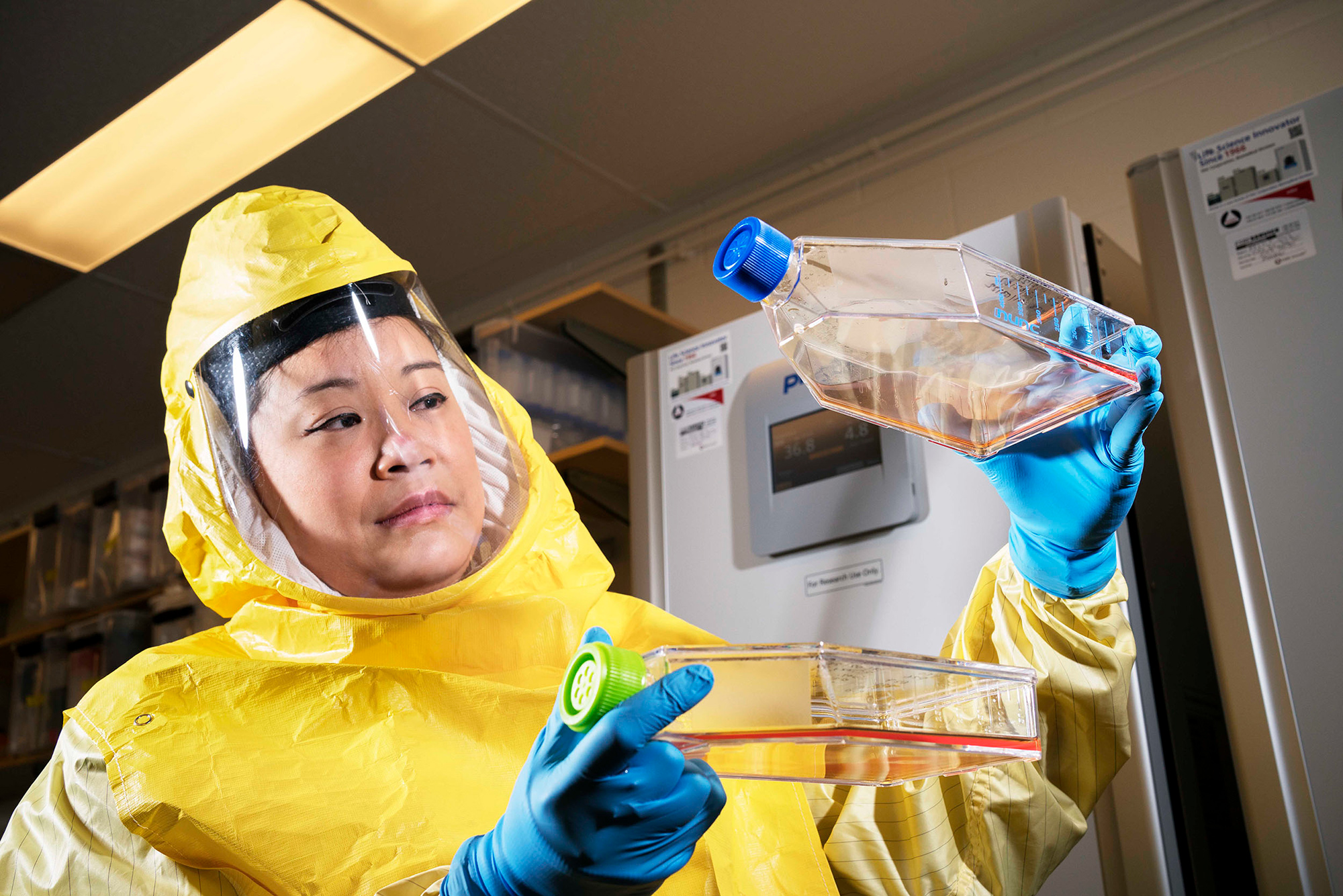Jessica Lam wearing a yellow hazmat suit, holding up and examining two flasks containing different coloured liquids