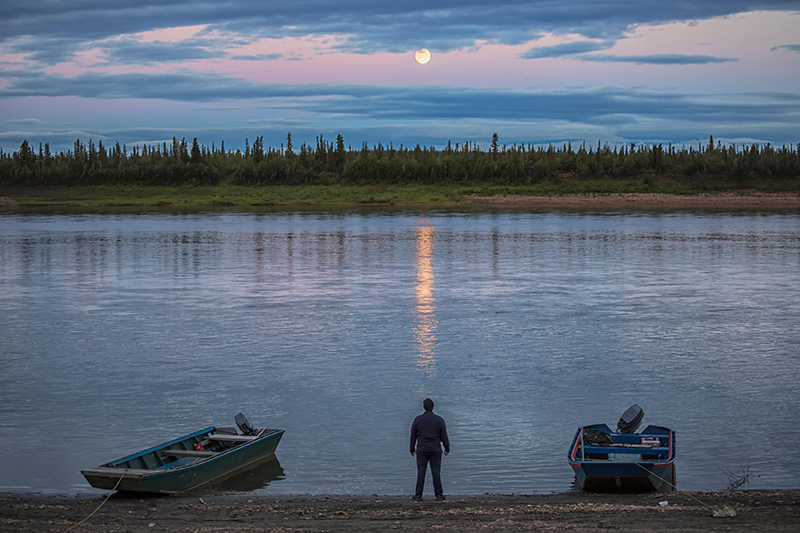 A man standing between two boats, looking across a river at the full moon in the background