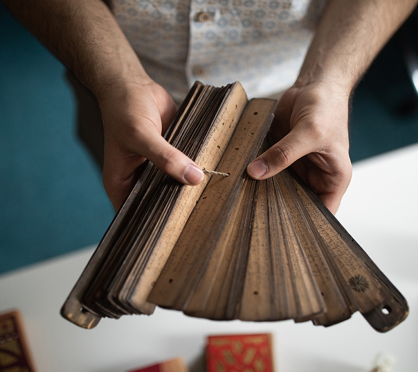 A pair of hands open the ruler-shaped pages of a Thai palm leaf manuscript like a fan