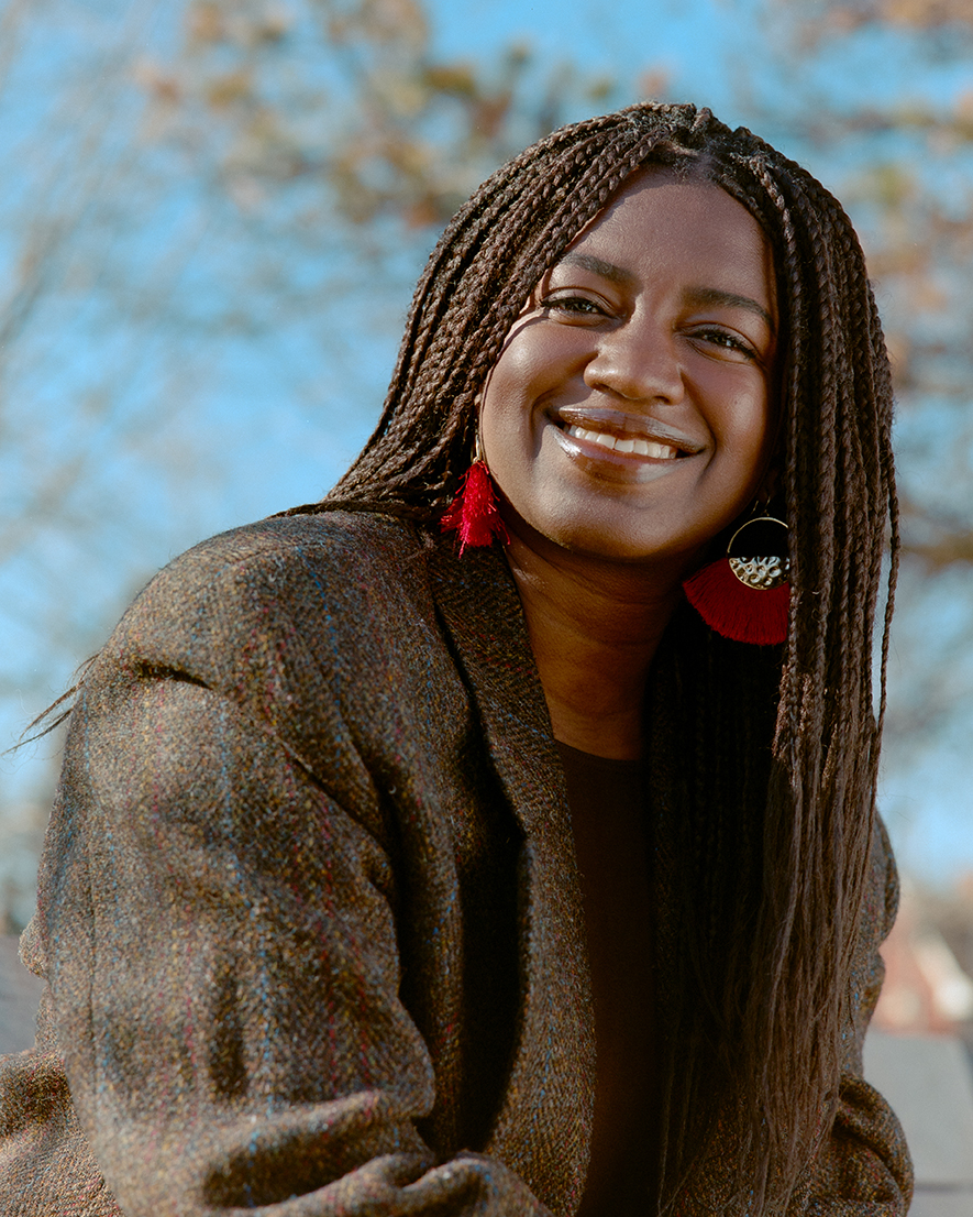Outdoor close up of Téa Mutonji, wearing a brown tweed coat and red and silver hooped earrings, smiling at the camera on a sunny day