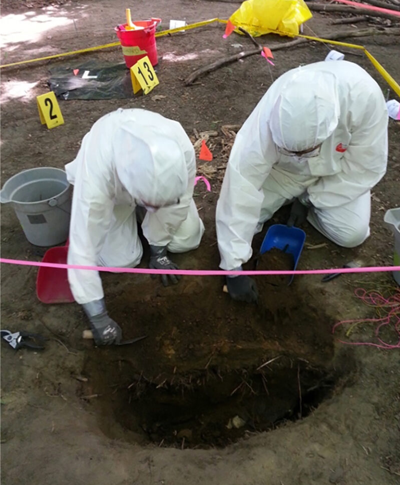 Two students in white forensic suits digging a hole in the ground in a cordoned off area