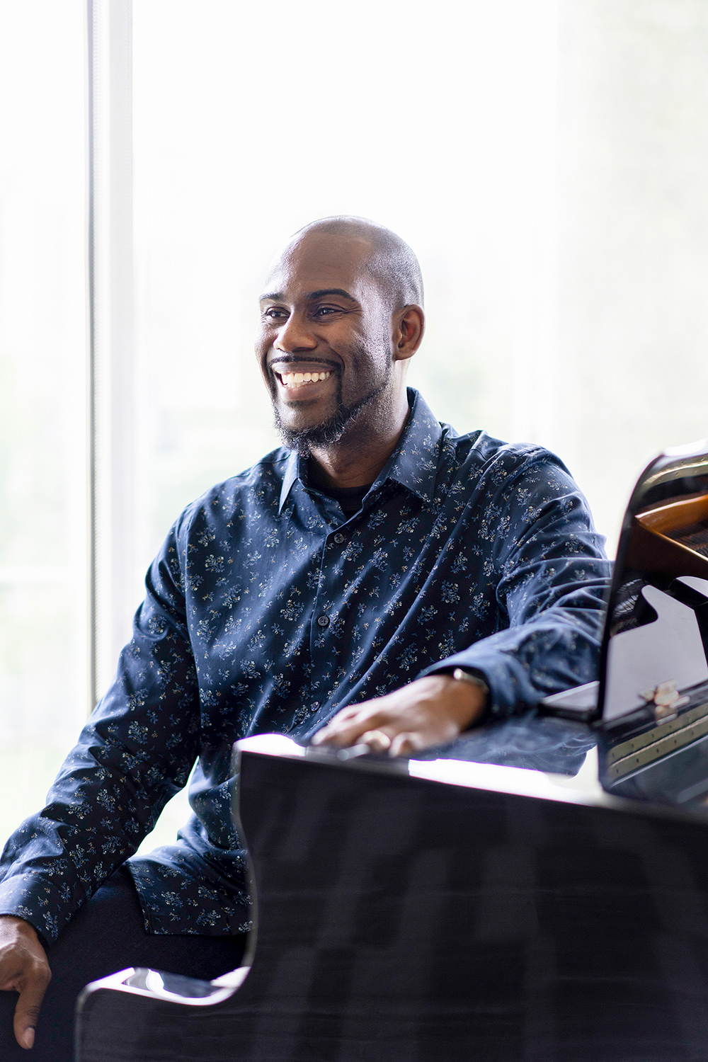 Darren Hamilton in a blue patterned, long-sleeved shirt, seated in front of a piano, smiling and looking off camera