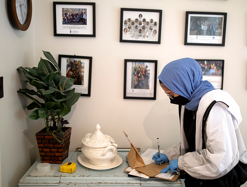 A student wearing a black face mask and blue hijab is writing on a brown paper evidence bag