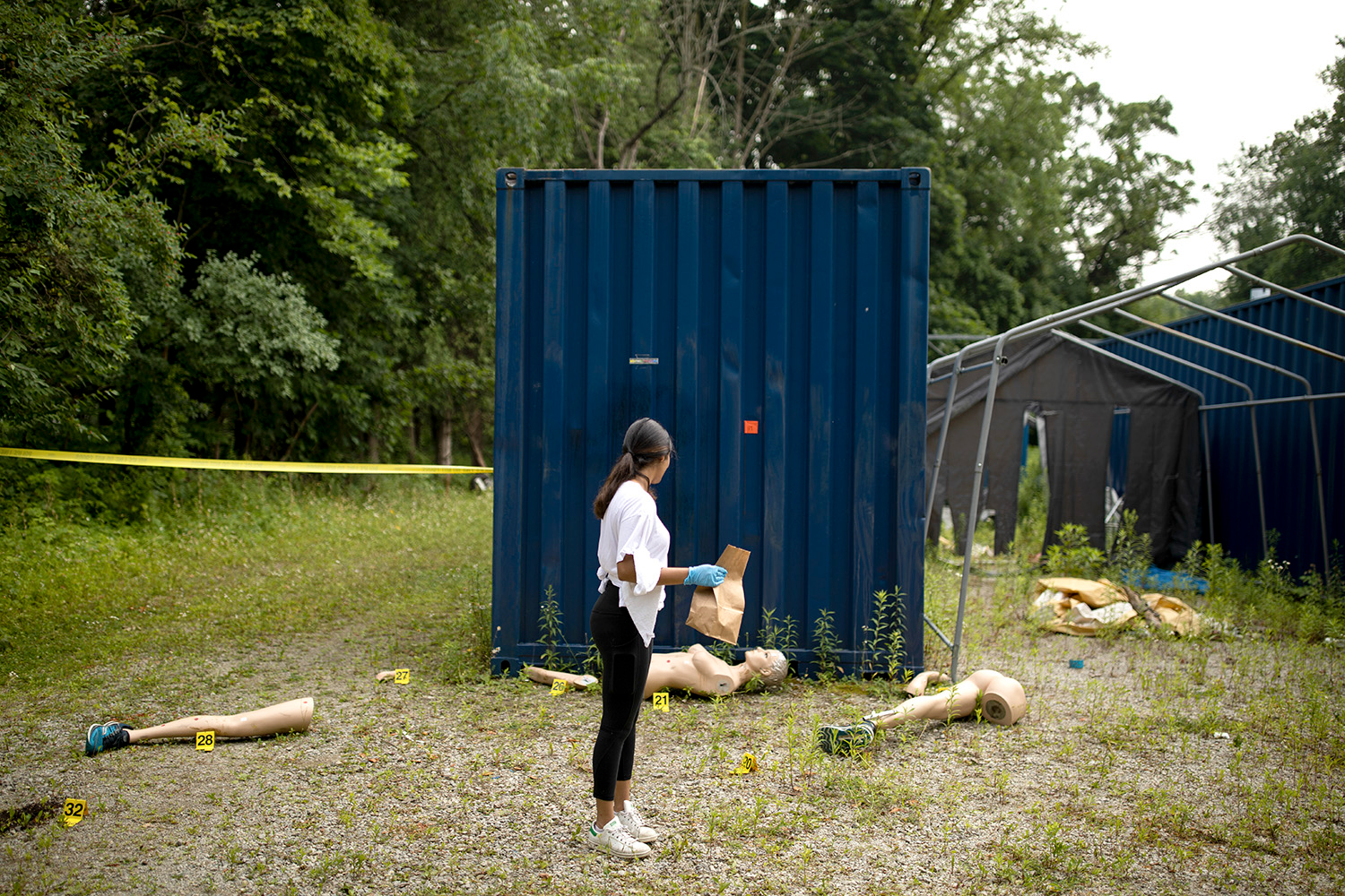 A student wearing blue gloves and holding a paper evidence bag looks back at the body of a female-shaped dummy lying in front of a large blue storage container, with its dismembered limbs scattered on the ground. Yellow labels marked with numbers are set beside each piece of evidence.
