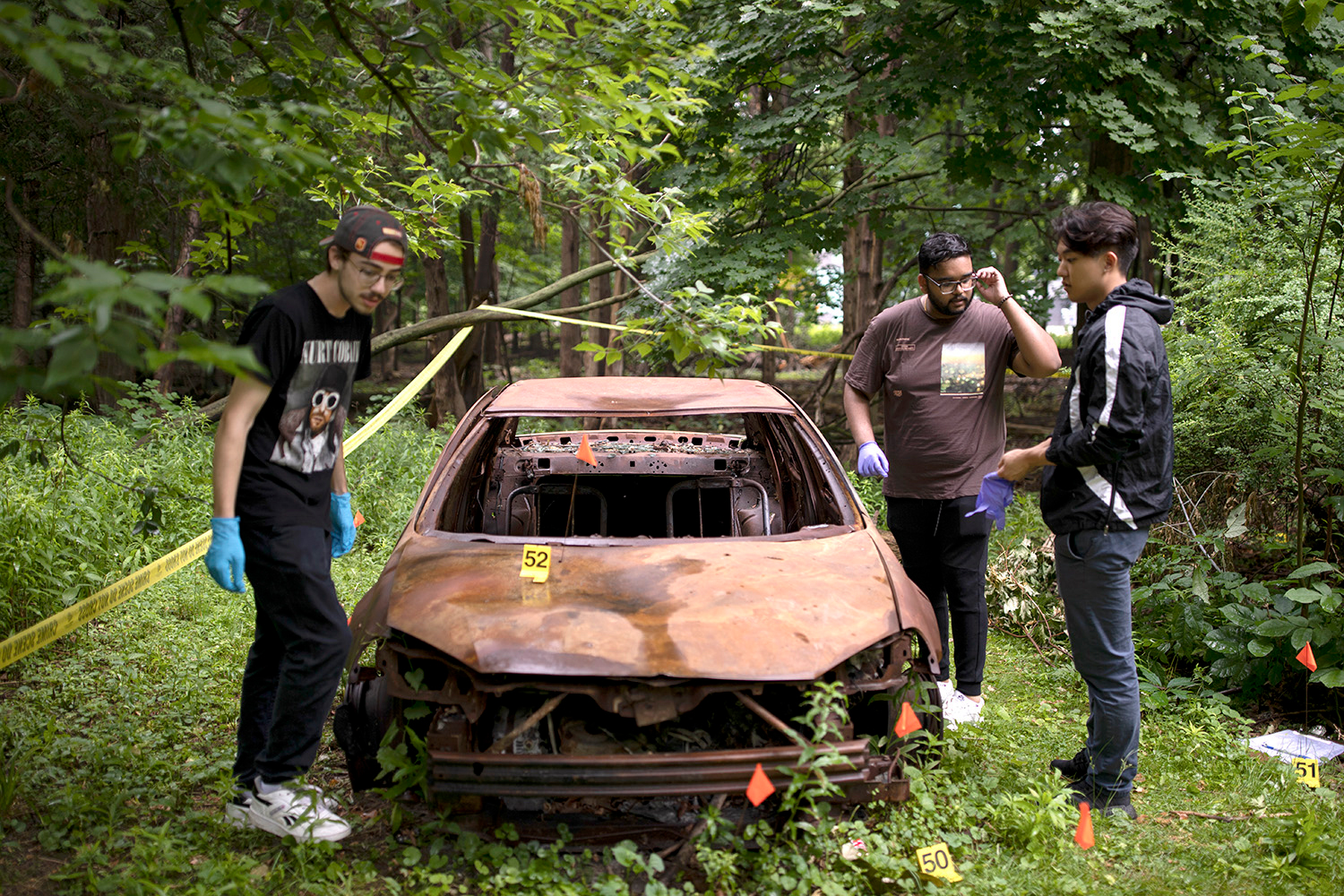 Three students wearing blue gloves examine a broken, rusted car in a forest area, marked 