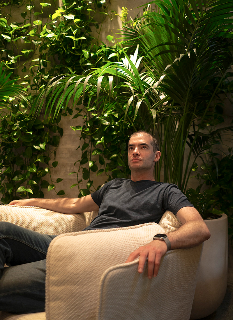Ilya Sutskever looks up while sitting on a beige lounge chair, with tall, leafy potted plants in the background