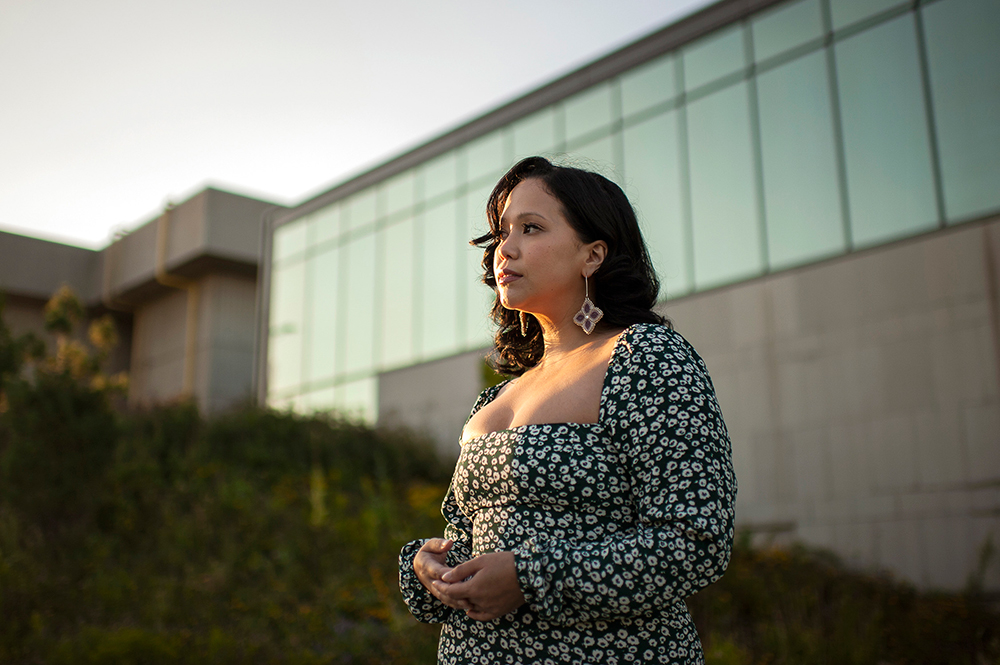 Prof. Kristen Bos wearing a long-sleeved, black and white flower patterned dress and large purple clover-shaped earrings, facing off camera, with a glass and concrete building and a grassy hill in the background