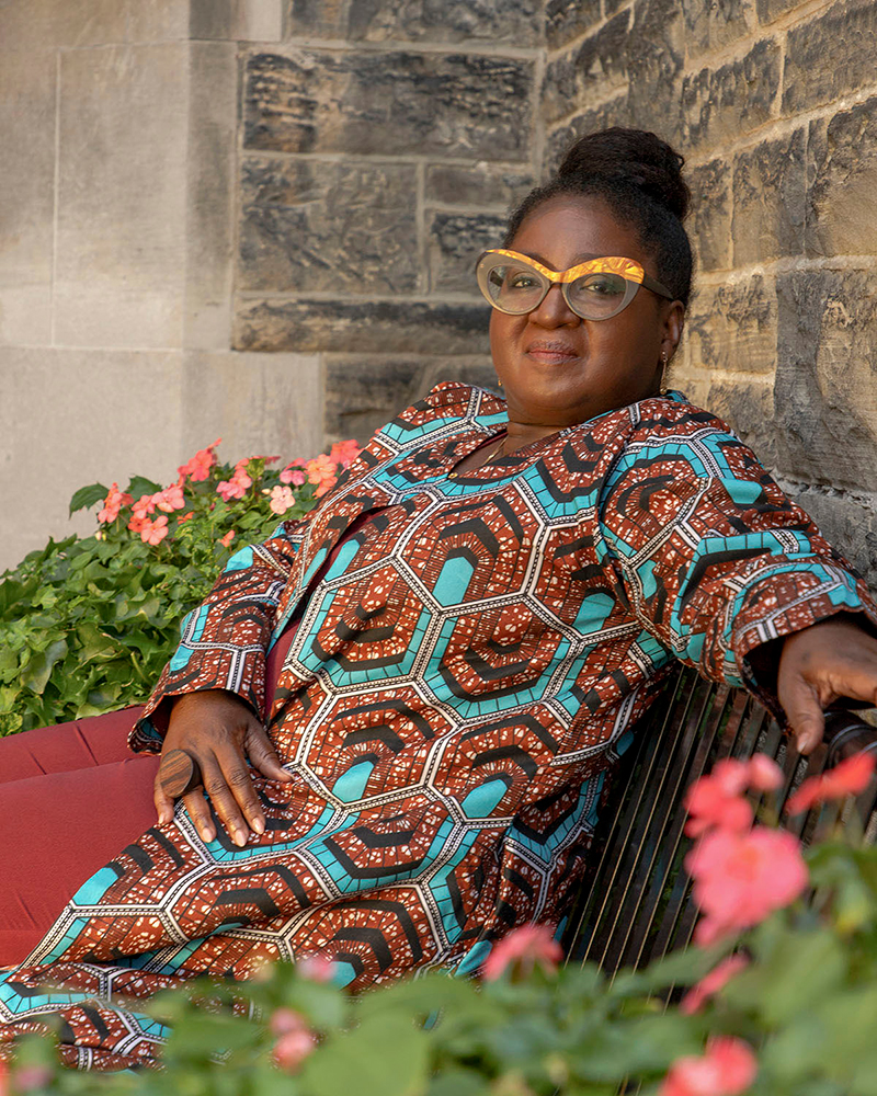 Prof. Notisha Massaquoi, wearing a maroon and teal hexagon-patterned tunic and yellow rimmed glasses, sitting on a black metal bench outdoors