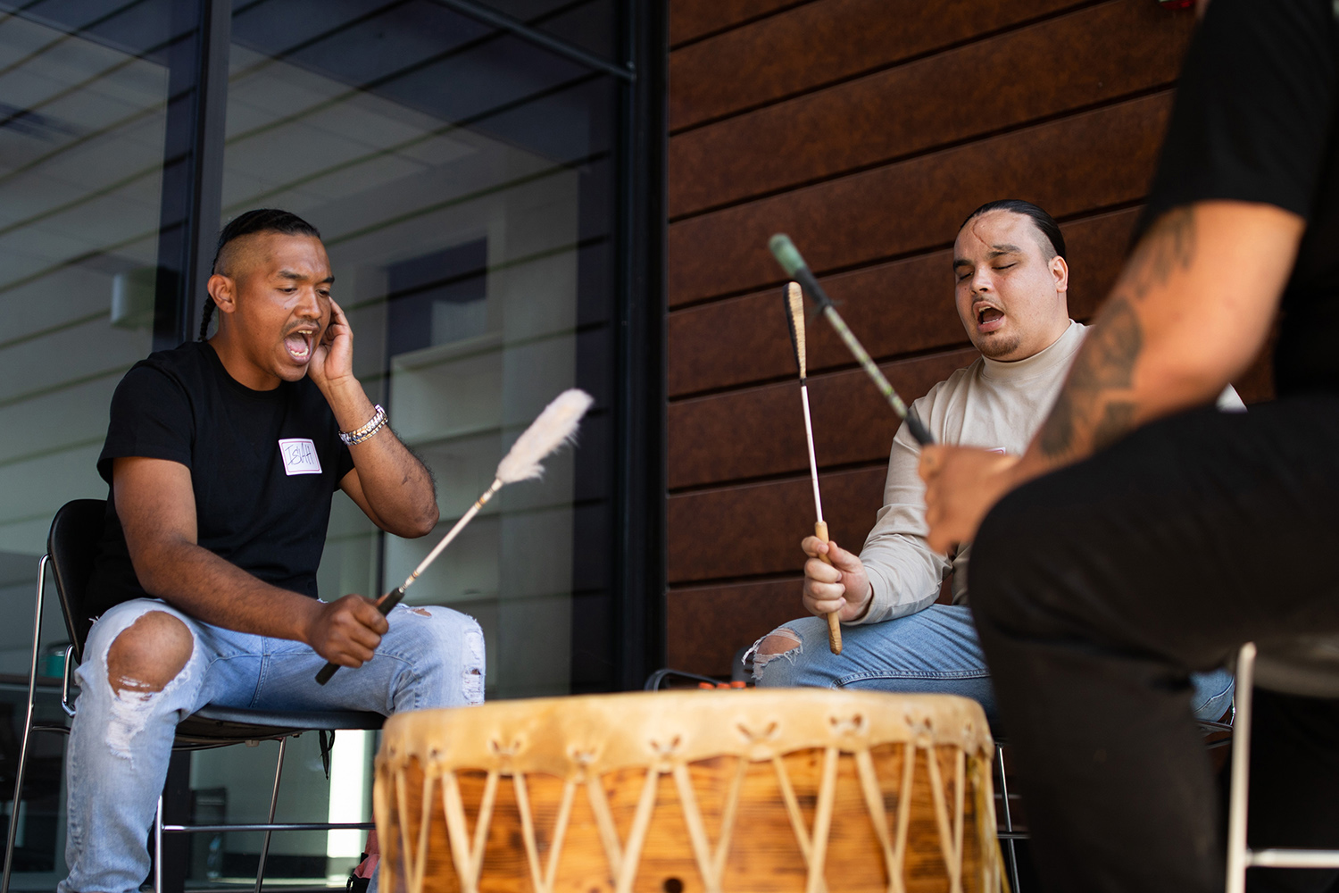Members of the Tsaunka Sugar Drum Group seated in a circle, performing on drums and singing.