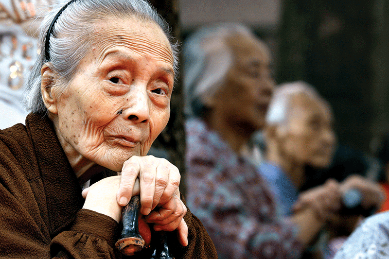 Close up photo of an elderly Chinese woman, sitting and leaning both hands on a cane, with two elderly people out of focus behind and to the right of her