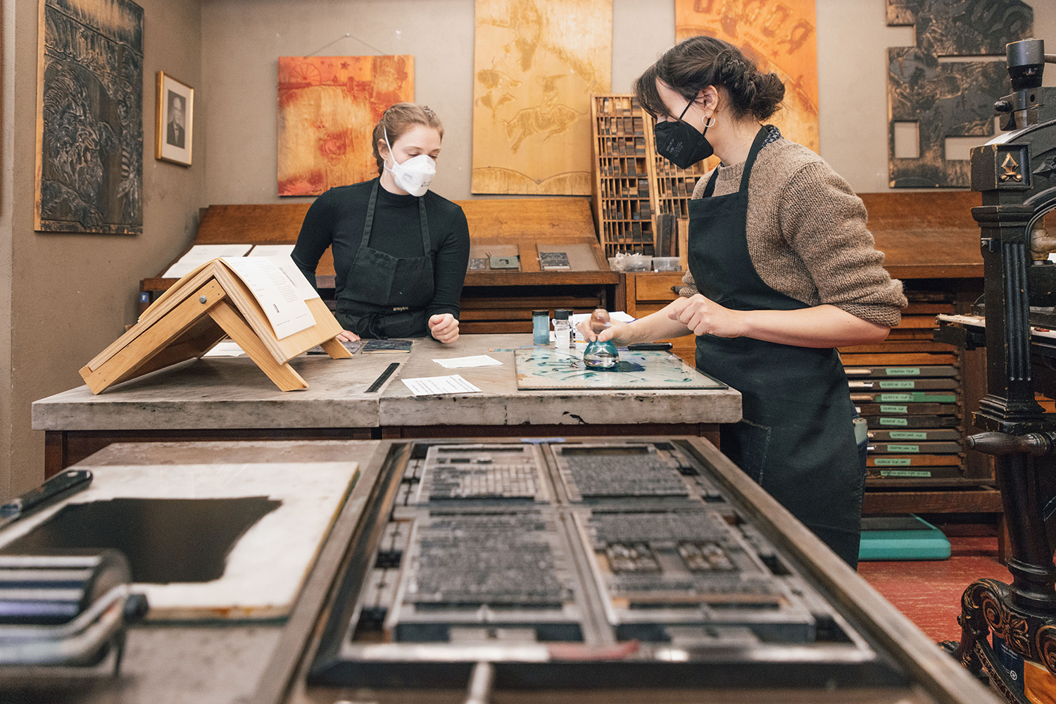 Two women stand at a desk. One is using a glass muller to blend green ink on the desktop as the other watches. In the foreground is a tray with type for letterpress printing