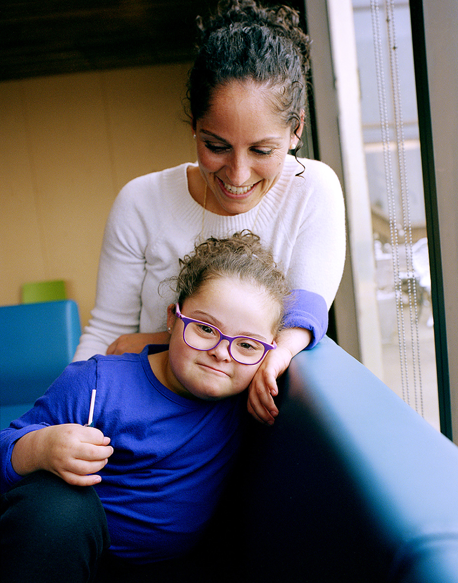 Prof. Christina Guzzo, looking down and smiling at her daughter, Stella, who is wearing purple-framed glasses. They are indoors, sitting on a blue bench.