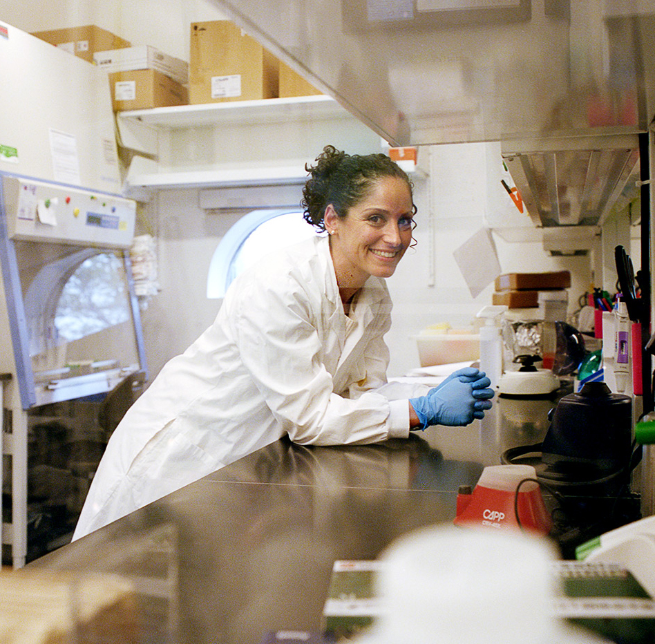 Prof. Christina Guzzo in a white lab coat and blue disposable gloves, leaning with her elbows against a lab table