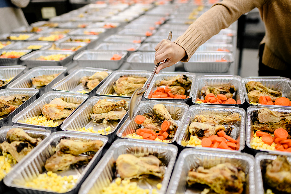 A volunteer spoons cooked carrots into aluminum food containers lined up in row upon rows, containing fried chicken legs and corn, some already containing carrots.