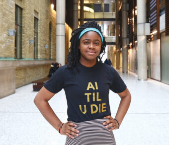 Inioluwa Deborah Raji, wearing a blue headband and a black T-shirt with the words "AI TIL U DIE" printed on the front, is standing with her hands on her hips in the front lobby of the Bahen Centre for Information Technology.