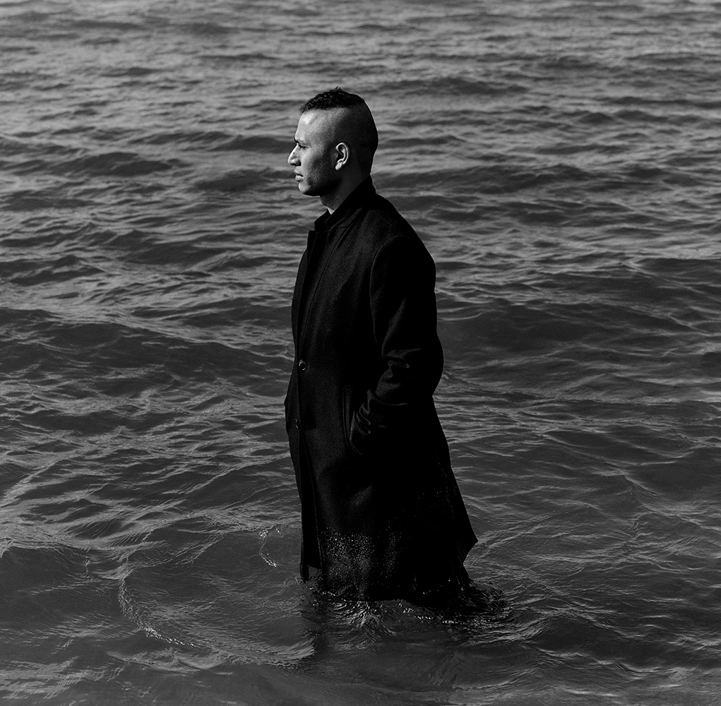 Black and white photo of Jiavet Ealom in a long black overcoat standing knee-deep in an open body of water with his hands in his pockets.