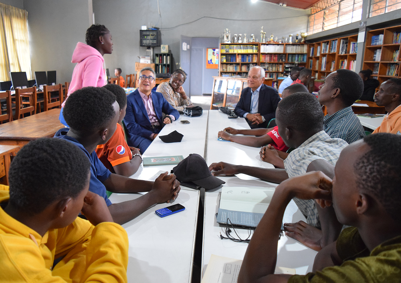 U of T President Meric Gertler and Prof. Joseph Wong, VP, International are sitting at a long table in a library with youth in the Giants of Africa program in Rwanda. One student, wearing a pink hoodie sweatshirt, is standing and talking with them.