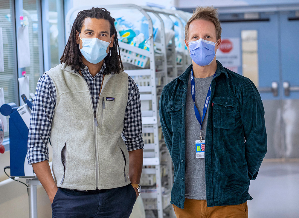 Dr. Mjaye Mazwi and Prof. Sebastian Goodfellow, standing side-by-side in a hospital hallway and wearing surgical facemasks