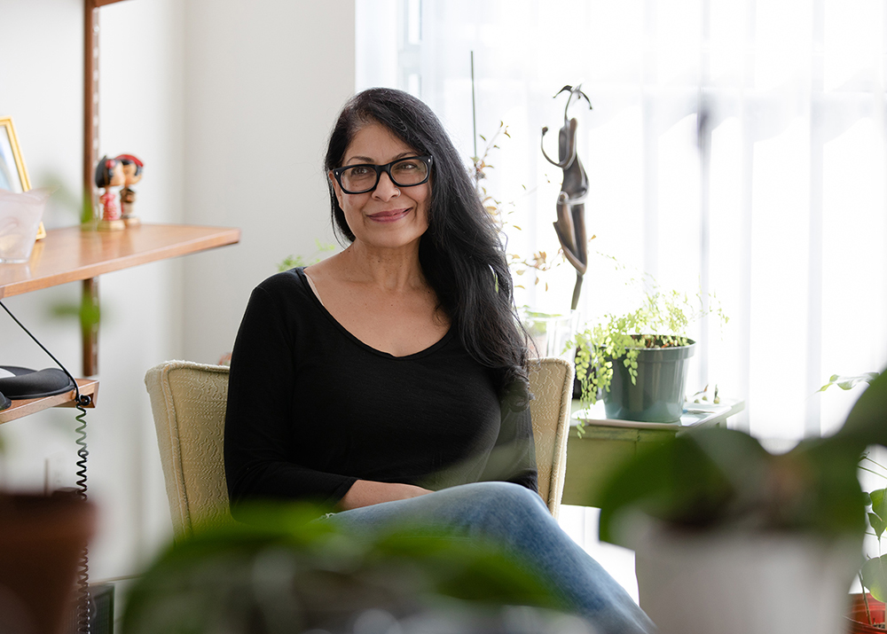 Nisha Pahuja, wearing a black 3/4 sleeve top and black-framed glasses, sitting on a chair at a corner next to potted plants in front of large windows, with sunlight flooding the room