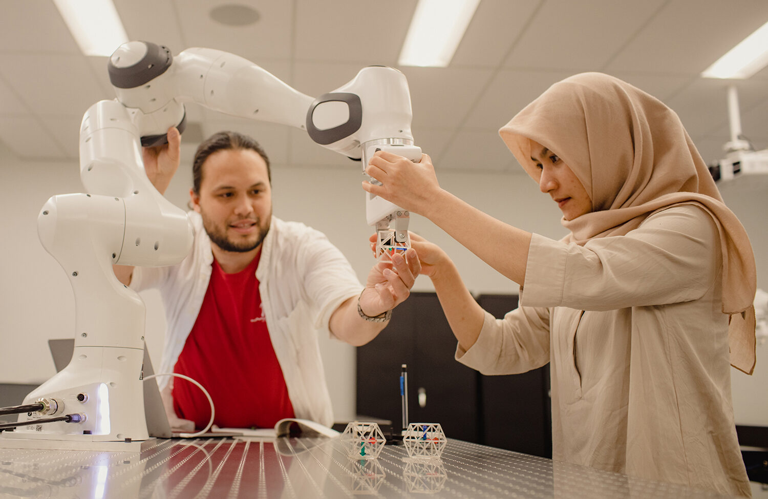 Reinhard Grassmann is holding a robotic arm steady with one hand and a plastic block under the grip of the cobot with the other, while Puspita Dewi, who is wearing a hijab, is adjusting the cobot's grip on the block.