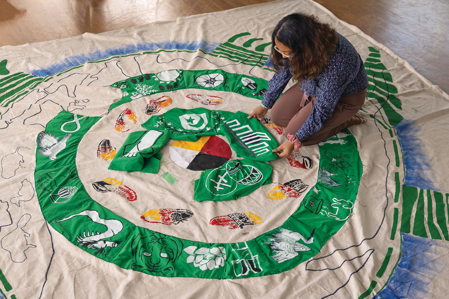 A cloth mural, depicting symbols sewn around a green outer ring as well as green inner ring, on top of the back of a turtle, with a medicine wheel in the centre, is spread out on the ground. Mandy Nelson is applying finishing touches on the inner ring of the mural.