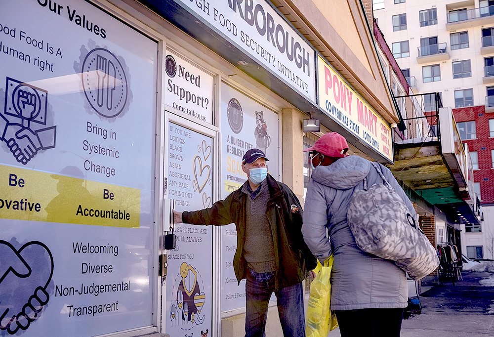 A volunteer, wearing a facemask with a hand on the door to the Scarborough Food Bank, greets and welcomes a client.