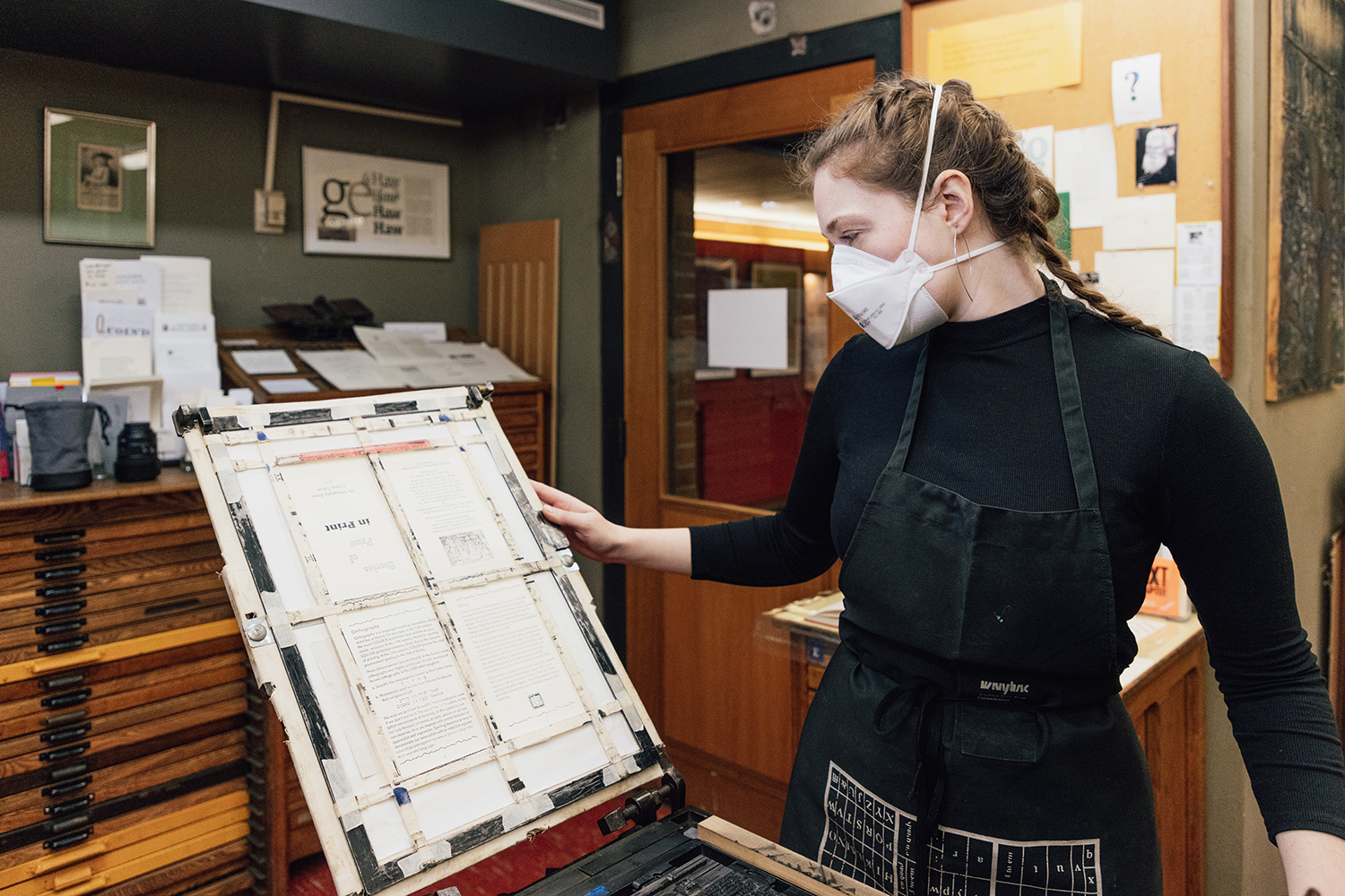 A woman looks at a page she has just printed. The page is attached to the tympan part of an iron hand press. The woman is wearing a face mask over her mouth and nose to protect against COVID-19.