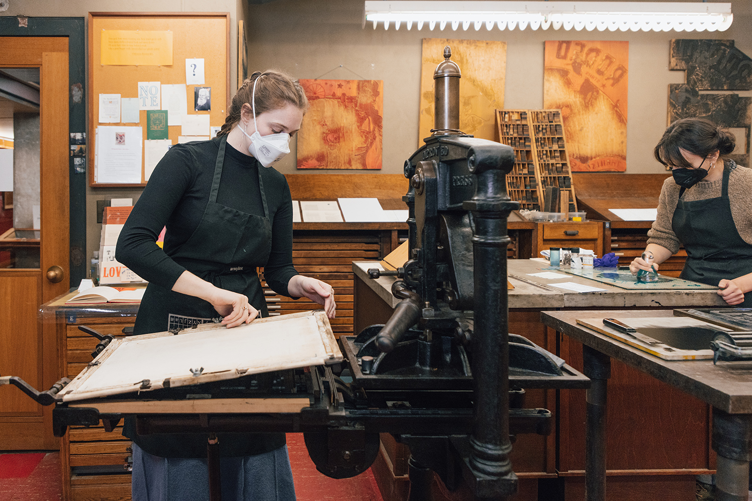 A woman covers a tray of type as she prepares to use a 19th century iron hand press. Another woman in the background works at a desk, making her own green ink.